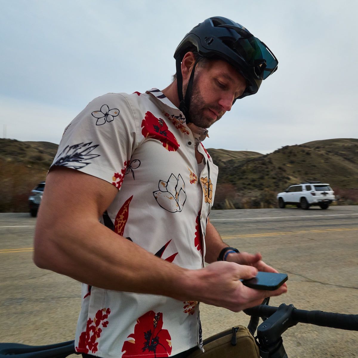 Man on a bicycle wearing a helmet and floral shirt, using a phone with a desert landscape in the background.
